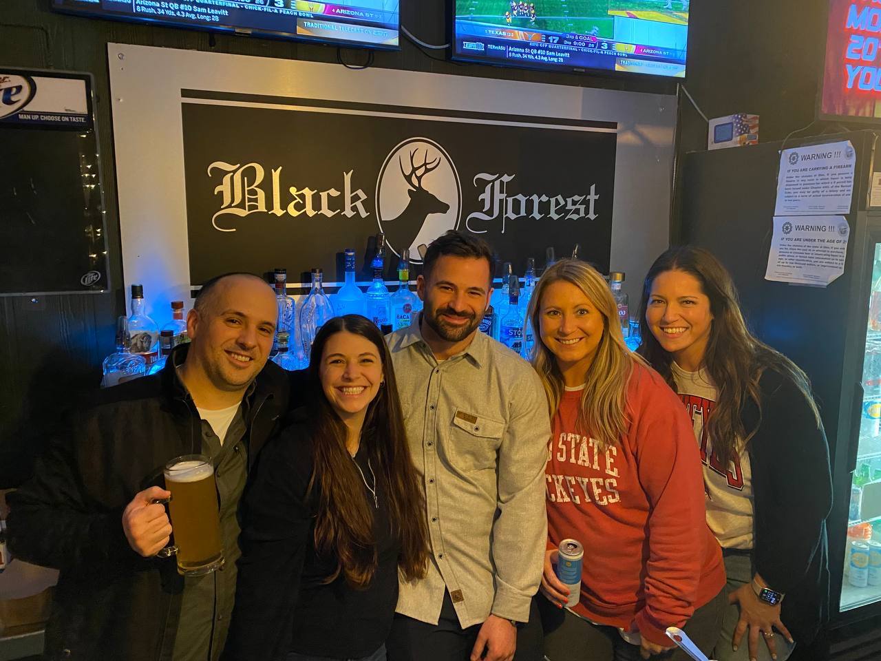 Five friends smiling together at the bar under the Black Forest sign